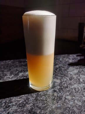 Narrow glass filled with a golden cocktail, the top half of which is white froth
